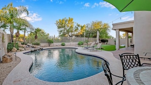 Piscina com borda curva, cercada por um espaço de terraço com espreguiçadeiras e uma área de estar coberta.