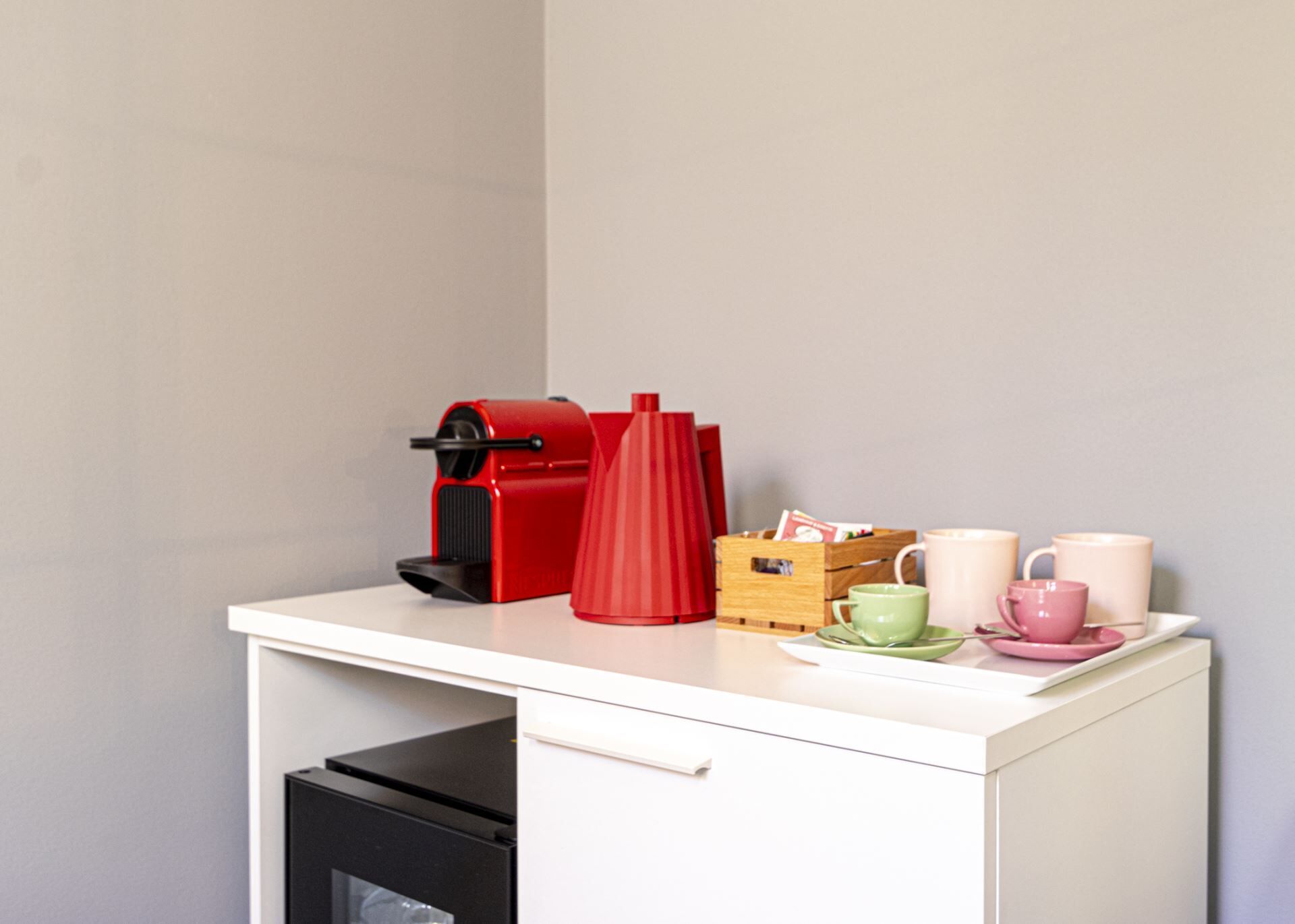Eine Küchenzeile mit einer Kaffeemaschine, einer roten Vase, einer Holzkiste und zwei Tassen auf einer weißen Arbeitsplatte.