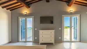 A spacious room with a high ceiling, a flat-screen TV mounted on the wall, and a white dresser. There are two sliding glass doors leading to a balcony with a view of the sea.