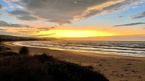A beach with a curved shoreline, grassy dunes, and a sunset over the ocean.