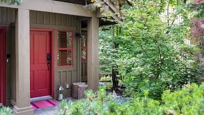 A wooden porch with a red door and shutters, surrounded by lush greenery and trees. - Modern Mountain Retreat | Ski-In | Hot Tub (Whistler)
