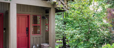 Une véranda en bois avec une porte rouge et des volets, entourée d’une végétation luxuriante et d’arbres.