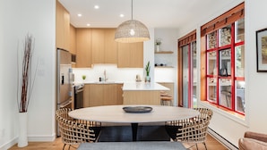 A modern kitchen with a central island, wooden cabinets, and a dining area with wicker chairs. - Modern Mountain Retreat | Ski-In | Hot Tub (Whistler)