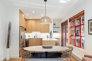 A modern kitchen with a central island, wooden cabinets, and a dining area with wicker chairs. - Modern Mountain Retreat | Ski-In | Hot Tub (Whistler)