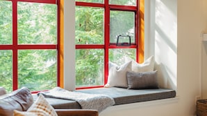 A cozy seating area with a bench and cushions, a window with red frames, and a view of greenery outside. - Modern Mountain Retreat | Ski-In | Hot Tub (Whistler)