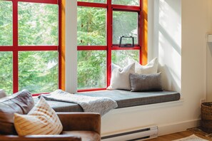 A cozy seating area with a bench and cushions, a window with red frames, and a view of greenery outside. - Modern Mountain Retreat | Ski-In | Hot Tub (Whistler)