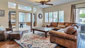 A spacious living room with a brown leather sofa, a wooden coffee table, and a patterned rug. A ceiling fan is visible, and there are large windows with blinds. - Private Pool, Pool Table, BBQ, Near San Diego Padres & Seattle Mariners Spring Training + FREE GOLF! (Glendale)