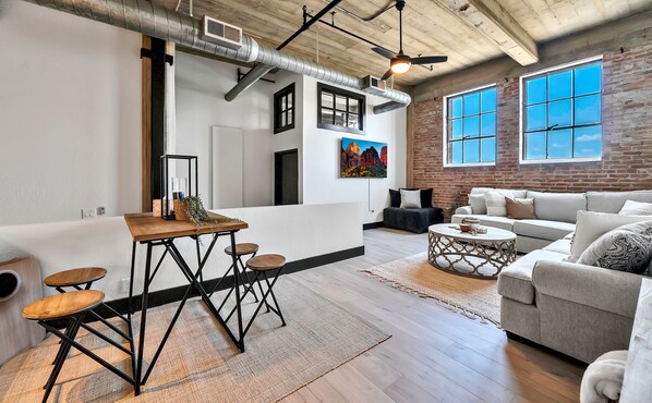 A spacious living area with a modern kitchen, a grey sofa, and a dining table with stools.