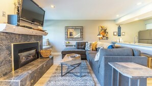 Un salón moderno con chimenea, un sofá gris, un televisor de pantalla plana y una cocina al fondo.