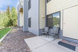 A building with a modern design, featuring a large window and a door. There are two chairs outside the building, and a small garden area with trees and a lawn.