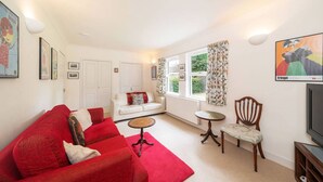 A living room with a red sofa, a white sofa, a small round table, and a wooden chair. There is a television on a stand and a window with curtains. - Earnside Cottage, Tranquil Escape in Comrie (Comrie)