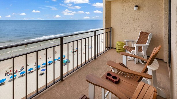 A balcony with a view of the beach, a wooden table, two wooden chairs, and a small table with a yellow stool. - Perfect 1 Bedroom Oceanfront!! (Murrells Inlet)