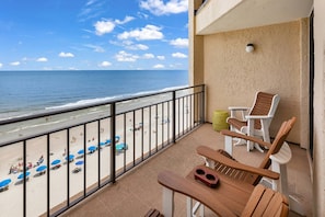 A balcony with a view of the beach, a wooden table, two wooden chairs, and a small table with a yellow stool.