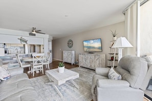 A spacious living room with a dining area, a TV mounted on the wall, and a ceiling fan.