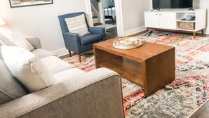 Un salon avec un canapé, une table basse, un fauteuil bleu, un téléviseur posé sur un meuble blanc et un tapis à motifs.