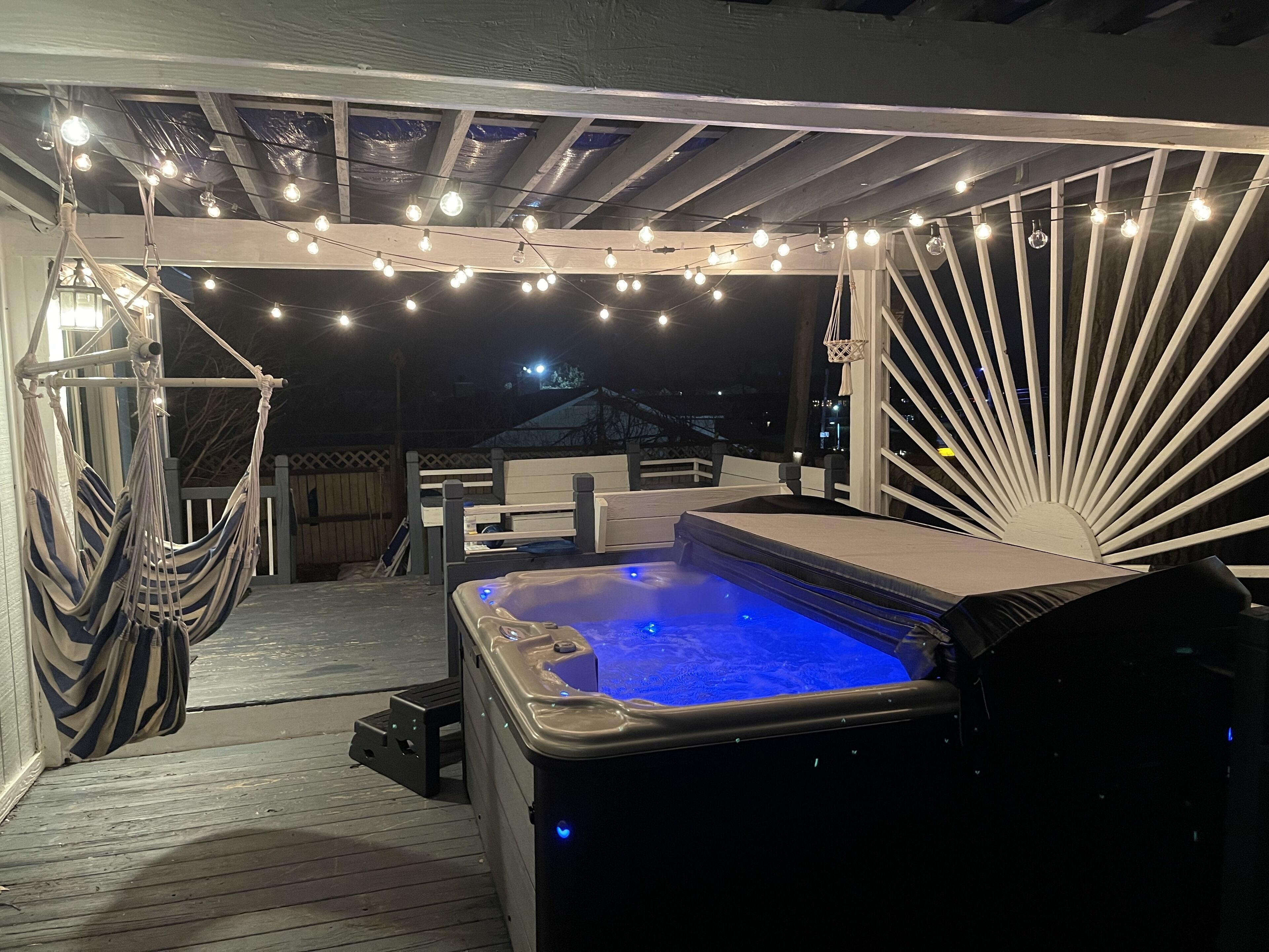 A hot tub under a pergola with string lights, surrounded by wooden deck and hammocks.