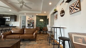Un salon moderne avec un canapé en cuir marron, une table à manger et des tabourets de bar. On y trouve un ventilateur de plafond, un coin cuisine avec un four à micro-ondes et une cuisinière, ainsi qu'un mur vert orné d'un néon.