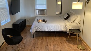 Une chambre à coucher comprenant un lit, une chaise, une table de chevet, une lampe, un téléviseur et une fenêtre avec des stores.