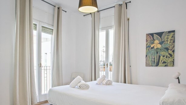 A bedroom with a bed, curtains, a painting, and a view through the window. - Duplex en Las Ramblas: tu refugio en BCN (Barcelona)