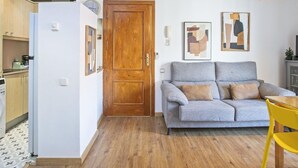 A compact living space with a kitchenette, a grey sofa with cushions, a wooden door, and two abstract paintings on the wall. - Duplex en Las Ramblas: tu refugio en BCN (Barcelona)