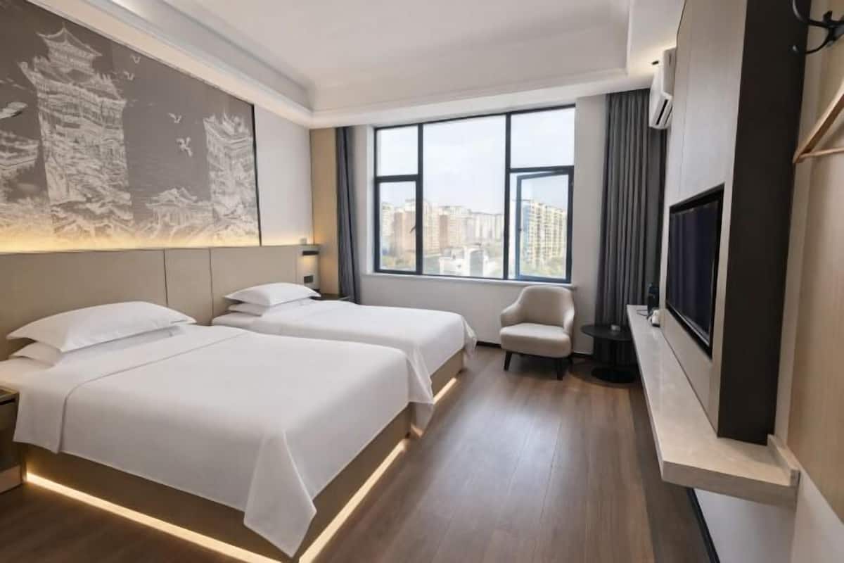 A hotel room with two beds, a chair, a TV, and a large window with a city view.