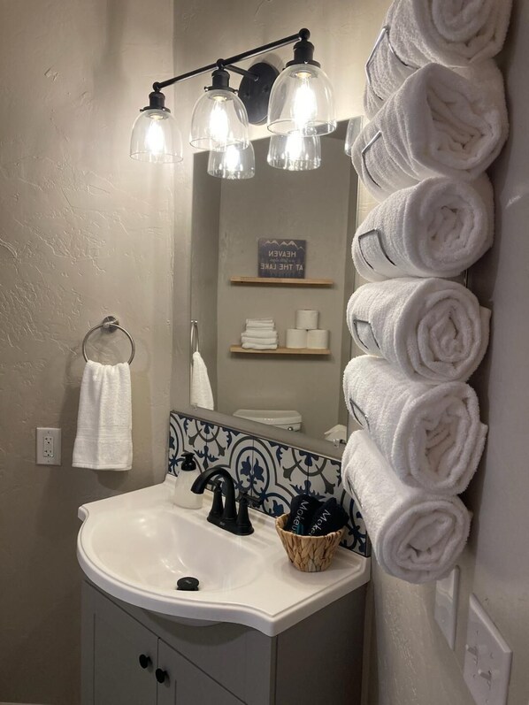 A bathroom with a white sink, black faucet, and a mirror with a towel rack. There are three light fixtures above the mirror and a shelf with towels and toilet paper.
