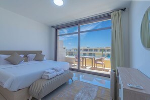 A hotel room with a large bed, a small desk, a balcony with a table and chairs, and a view of buildings. - Vesta - Luxury APT - 1BR - Gcribs I (El Gouna)