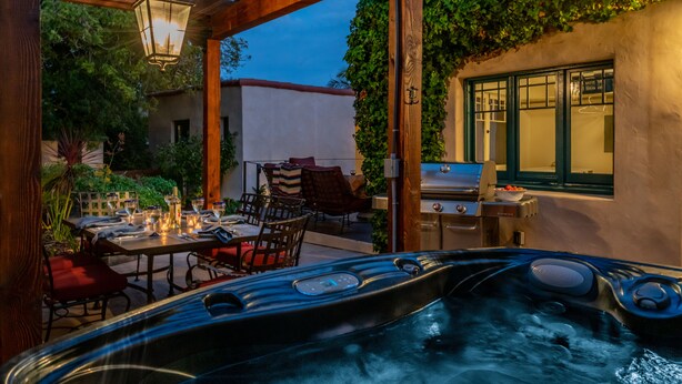 Un jacuzzi bajo una pérgola de madera, con una mesa y sillas cerca, rodeado de vegetación y vistas a un edificio.