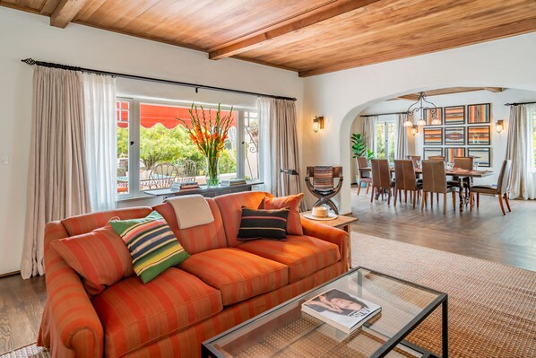 A living room with a wooden ceiling, a sofa with colorful cushions, a glass coffee table, and a dining area with a wooden table and chairs. - Historic Adobe Retreat | Firepit Courtyard, Private Hot Tub & Timeless Character (Santa Barbara)