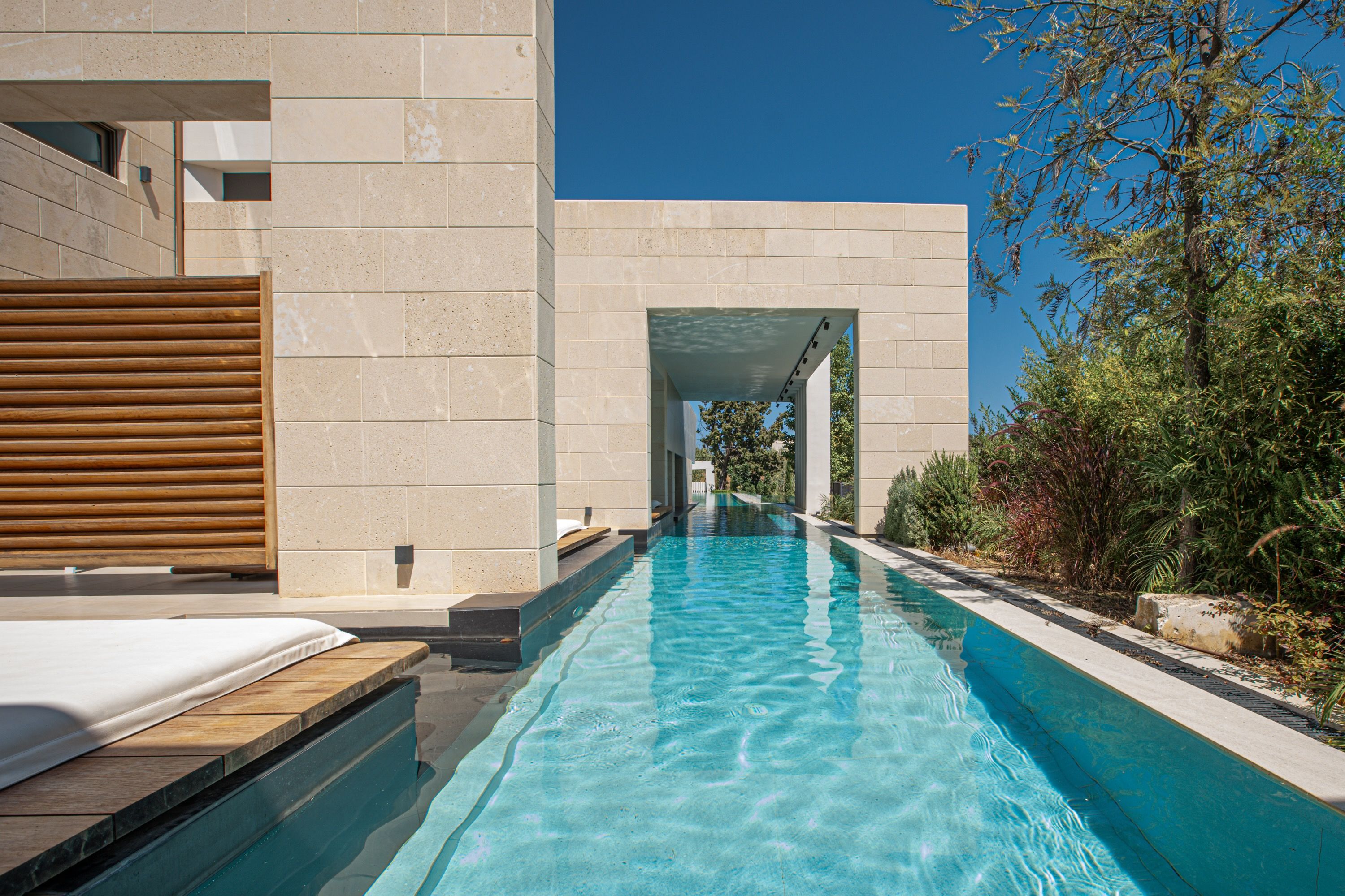 A swimming pool with a clear blue water surface, surrounded by a modern building with beige stone walls and a wooden deck.