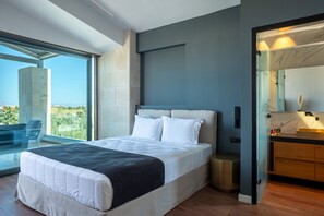 A modern bedroom with a large bed, a view of the outdoors, a bathroom with a mirror, and a small seating area.