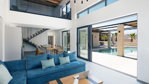 A modern living room with a blue sofa, a wooden coffee table, and a laptop. There is a staircase leading to an upper level, and a pool area is visible through large windows. - Ikritis Villa Ι - 1,2Km to beach & sea views! (Chania)