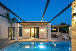 Ein moderner Poolbereich im Freien mit einer Steinmauer, einer überdachten Sitzgelegenheit und einem Eingang mit Glastür.
