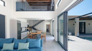A modern interior with a blue sofa, a kitchen, a dining area, and a glass-enclosed outdoor space. - Ikritis Villa Ι - 1,2Km to beach & sea views! (Chania)