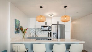 A modern kitchen with white cabinets, a stainless steel refrigerator, a microwave, and a coffee maker. There are two hanging wicker pendant lights, a blue tiled backsplash, and a white countertop.