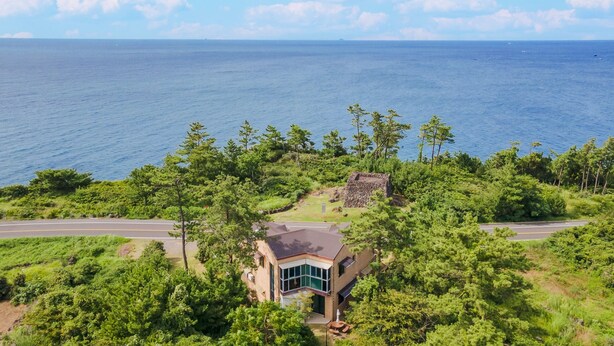 A coastal house with a large bay window, surrounded by lush greenery and a view of the ocean.