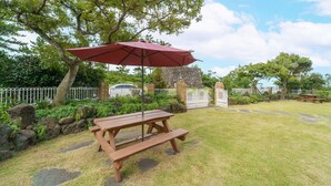 A picnic table with a red umbrella in a grassy backyard with a stone wall and white fence.
