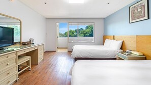 A hotel room with a large TV, wooden desk, two beds, a nightstand, and a window with a view of trees. - Jeju Honeycrown Hotel (Jeju City)