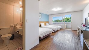 A hotel room with a wooden floor, a bed with white bedding, a window with a view of greenery, and a bathroom with a toilet and sink. - Jeju Honeycrown Hotel (Jeju City)