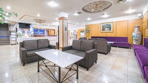 Un hall d’hôtel avec des canapés gris, une table blanche et un comptoir de réception portant les inscriptions « CAISSE » et « ENREGISTREMENT ».