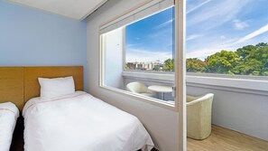 A hotel room with two beds, a large window with a city view, and a small balcony with a chair and table. - Jeju Honeycrown Hotel (Jeju City)