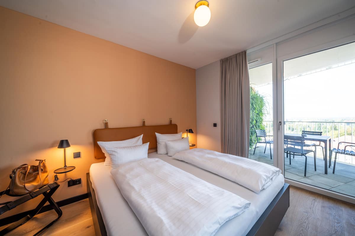 Ein Hotelzimmer mit zwei Betten, einem Nachttisch mit Lampe, einem Sessel und Blick auf den Balkon.