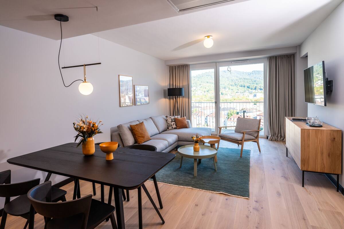 Ein modernes Wohnzimmer mit einer Couch, einem Esstisch und Stühlen. Ein Balkon bietet Blick auf die Hügel.