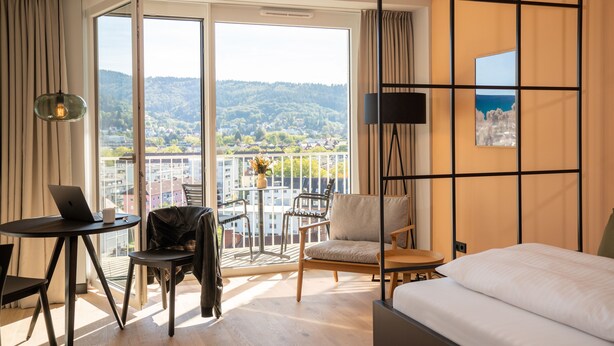Une chambre d’hôtel avec un lit, un bureau avec un ordinateur portable, une chaise, une table avec un vase et une vue sur la ville et les montagnes.