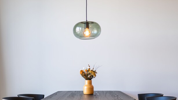 Un coin repas avec une table ronde, quatre chaises noires, un vase de fleurs et un luminaire suspendu.