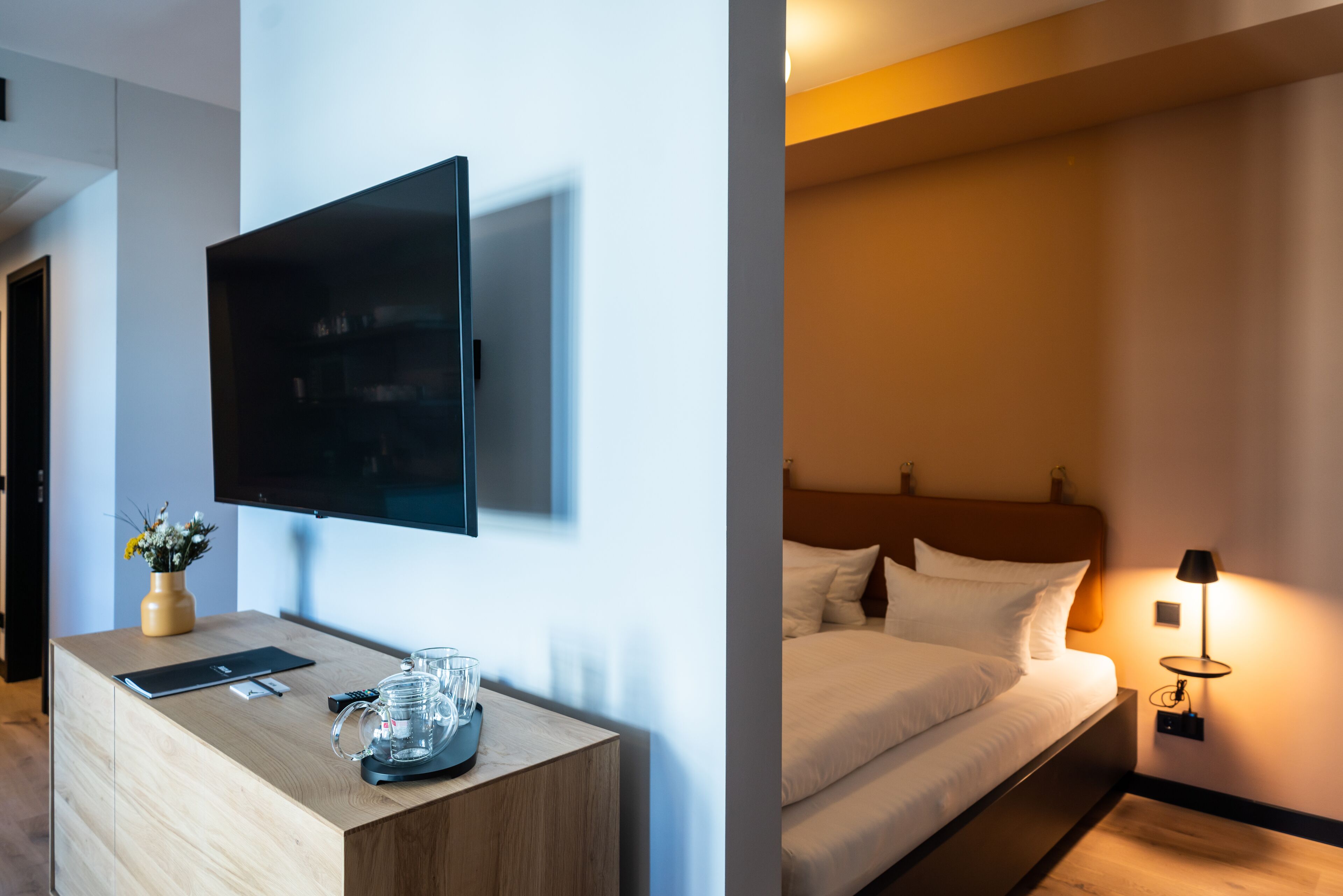 A hotel room with a flat-screen TV mounted on the wall, a wooden bedside table with a vase of flowers, a bed with white bedding, and a bedside lamp.