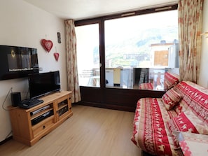 A room with a TV on a wooden stand, a bed with patterned bedding, and a large window with a view of buildings and mountains.