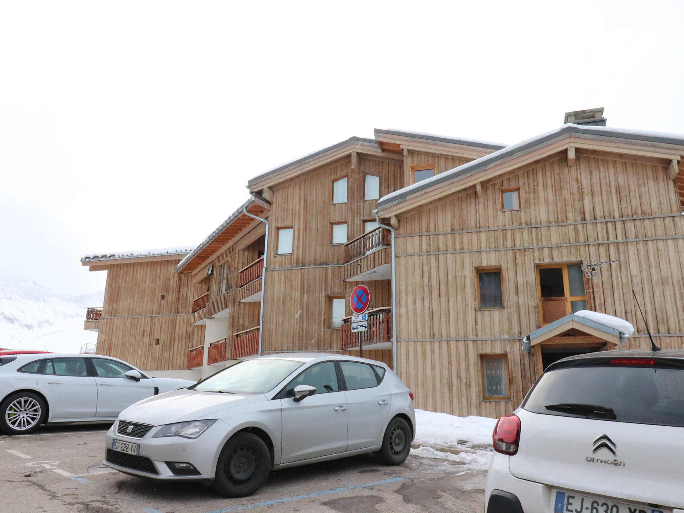A multi-story wooden building with balconies, a parking area, and a snowy mountain backdrop.