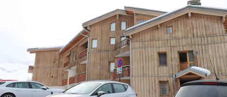A multi-story wooden building with balconies, a parking area, and a snowy mountain backdrop.