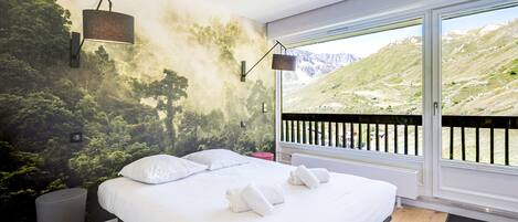 Une chambre dotée d’un grand lit, offrant une vue sur un paysage montagneux grâce à une porte coulissante, et dont un mur est décoré d’une fresque représentant une forêt.